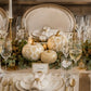 A styled dining table setting with white plates, gold-rimmed glassware, folded napkins, a place card reading 'Daniel', and decorative pumpkins with gold patterns on a green leafy runner, set against a beige upholstered chair.