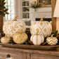 A decorative autumn pumpkin arrangement displayed on a wooden tray in a warm, elegant living room setting. The set includes multiple pumpkins in cream and gold patterns, including a large diamond-patterned pumpkin, a chevron-patterned pumpkin, and textured gold pumpkins, accented with green leaves.