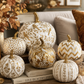 A styled collection of decorative pumpkins in white and gold, featuring various patterns like chevrons, dots, and floral motifs, arranged on a table with books, candles, and autumn decor.