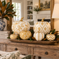 A styled autumn pumpkin decoration set in a wooden tray on a rustic wooden console table, featuring white and gold patterned pumpkins with green leaves, set in a beautifully decorated living room.