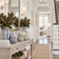 A styled entryway console table decorated with blue-and-white ceramic vases, patterned pumpkins, green foliage, books, and a lantern, set in a bright white hallway with a rug and staircase.