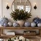 A styled home decor display featuring a collection of blue and white patterned pumpkins arranged on a rustic wooden console table in a bright room with a large round mirror and window views.