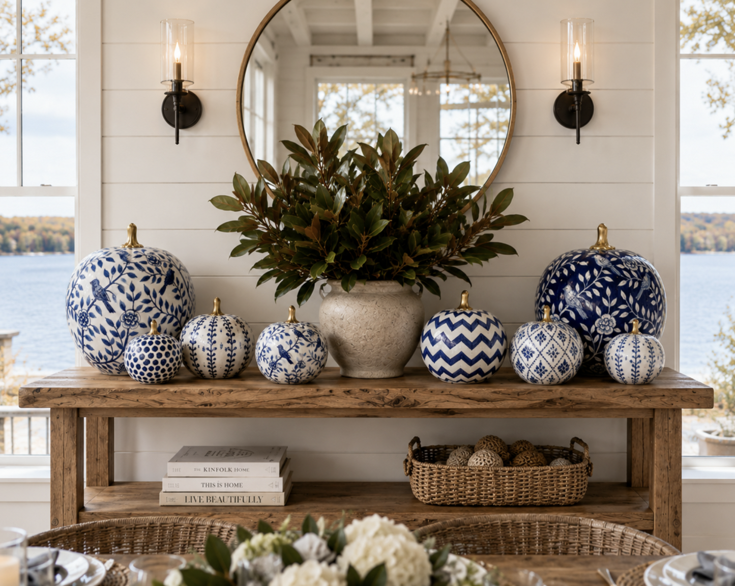 A styled home decor display featuring a collection of blue and white patterned pumpkins arranged on a rustic wooden console table in a bright room with a large round mirror and window views.