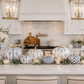 A styled kitchen island display featuring a collection of white and blue patterned pumpkins, white pumpkins, and tealight candles, set against a bright white kitchen background with marble backsplash and gold pendant lights.