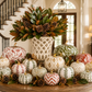 A styled autumn holiday centerpiece featuring a collection of patterned pumpkins in white, green, and red, arranged on a wooden table with pinecones and a large white lattice vase filled with green foliage and pinecones. The setting is a grand interior with a staircase in the background.