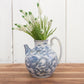 Blue and white patterned ceramic jug with greenery on a wooden surface and white wooden panel background