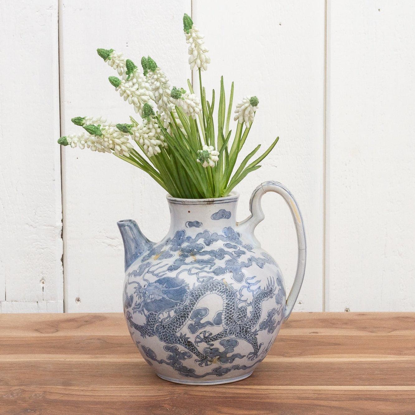 Blue and white patterned ceramic jug with greenery on a wooden surface and white wooden panel background