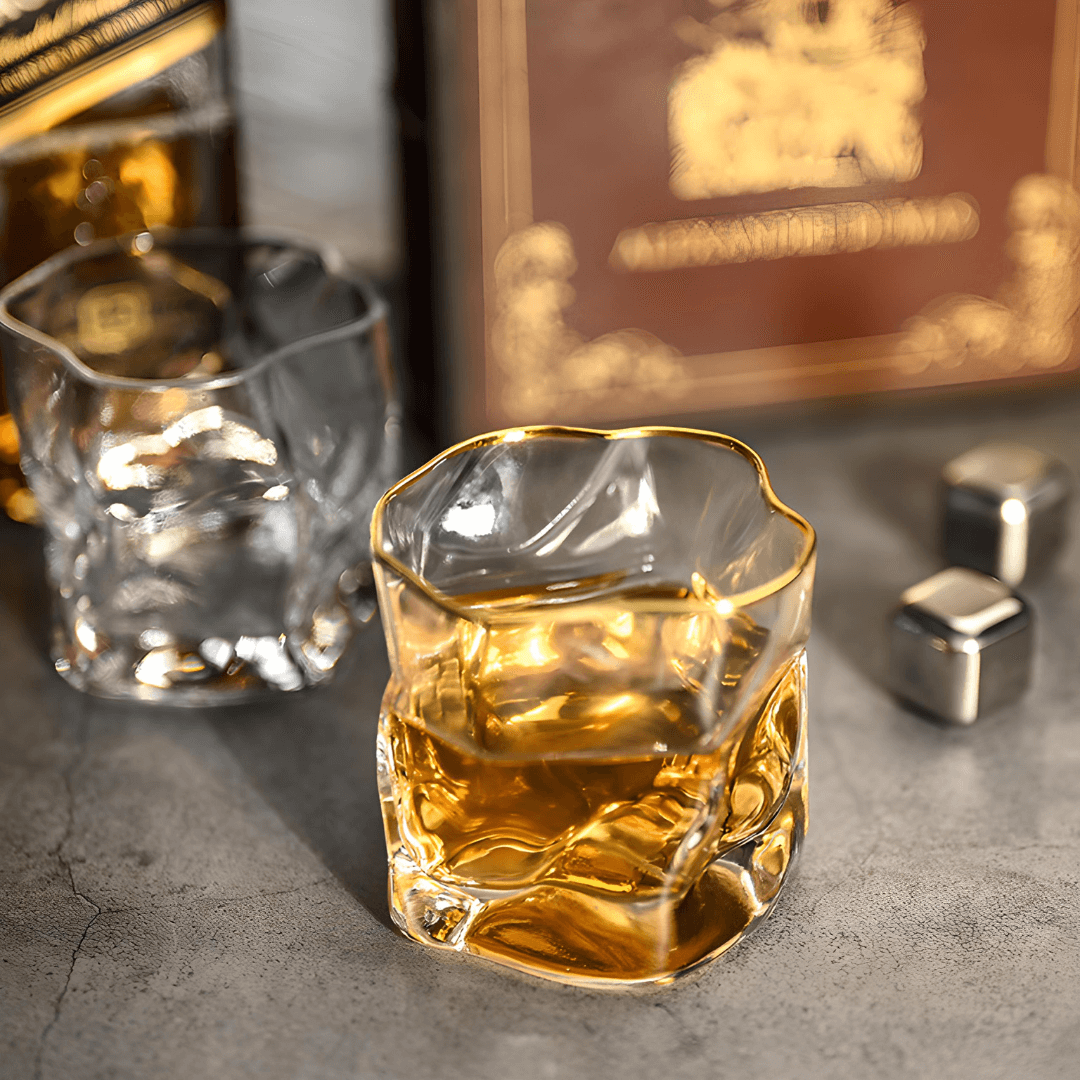 Glass of whiskey with ice cubes on a textured surface, with a blurred bottle in the background.