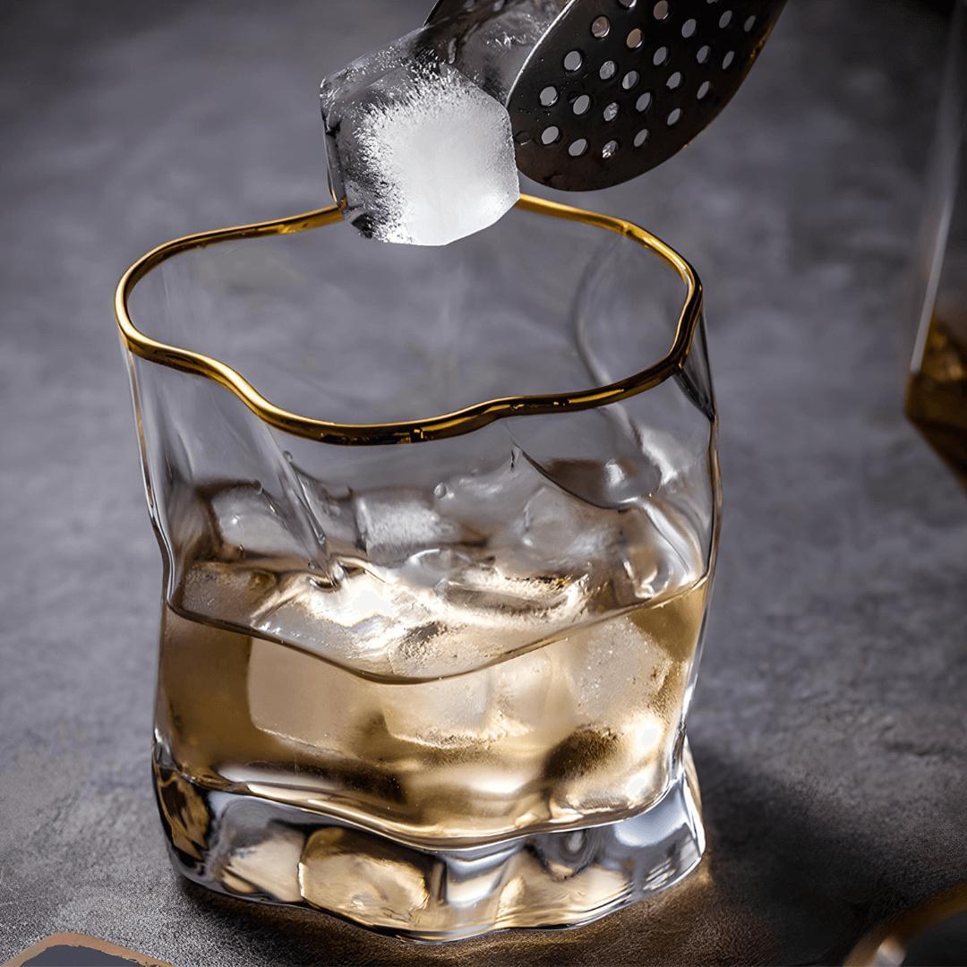 Glass of whiskey with ice being topped off with a gold-rimmed glass on a dark background