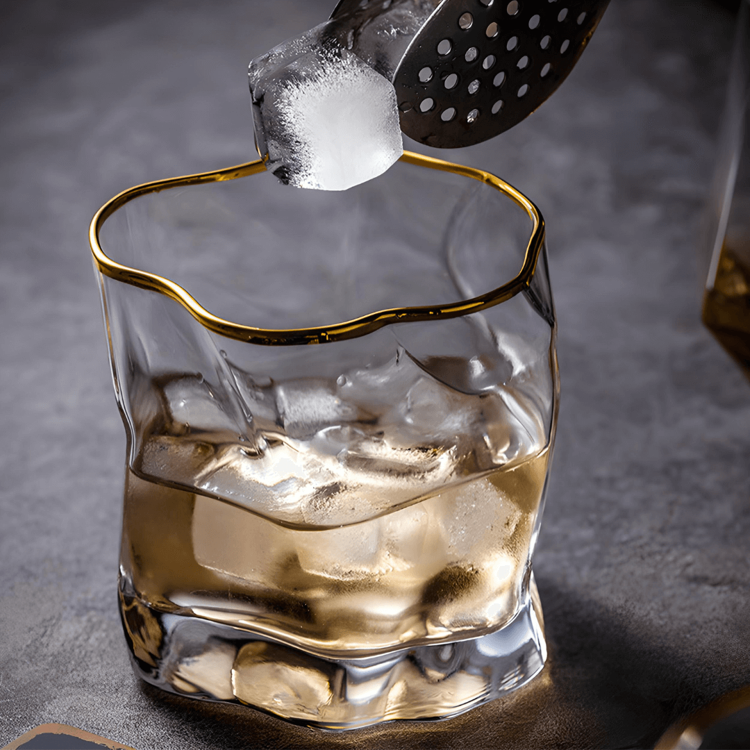 Glass of whiskey with ice being topped off with a gold-rimmed glass on a dark background