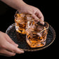 Two glasses of whiskey with ice on a black tray, held by two hands against a dark background.