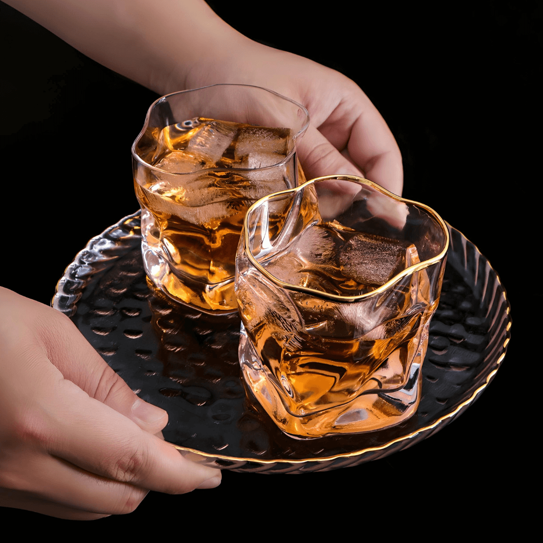 Two glasses of whiskey with ice on a black tray, held by two hands against a dark background.
