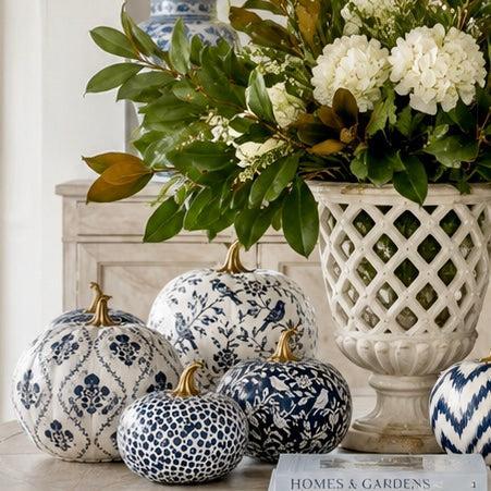 Decorative ceramic pumpkins with blue and white patterns arranged on a tabletop beside a large white lattice planter filled with green foliage and white flowers.