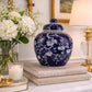 Decorative blue vase with white floral patterns on a table with books, flowers, and a lamp.