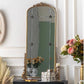 An ornate gold-framed floor mirror with a tall arched shape, standing on a dark console table in a decorated room with flowers and wall molding.