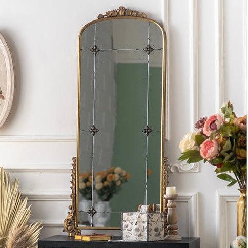 An ornate gold-framed floor mirror with a tall arched shape, standing on a dark console table in a decorated room with flowers and wall molding.