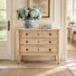 A light natural wood three-drawer chest of drawers with dark round knobs, styled in a bright room with a blue and white ceramic vase of flowers on top.