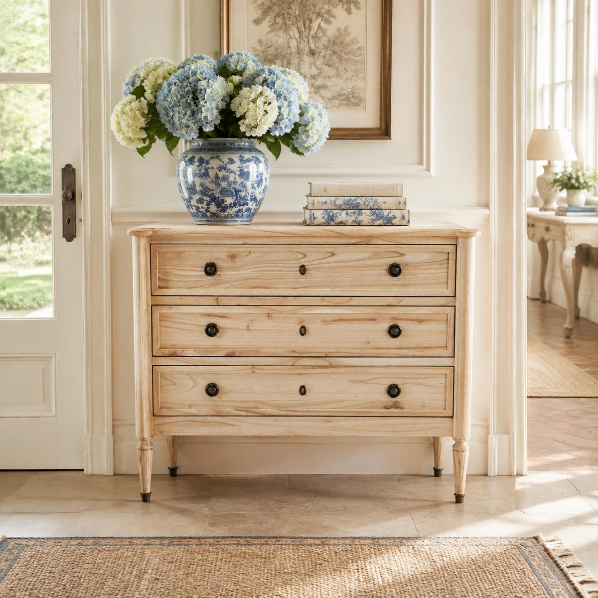 A light natural wood three-drawer chest of drawers with dark round knobs, styled in a bright room with a blue and white ceramic vase of flowers on top.
