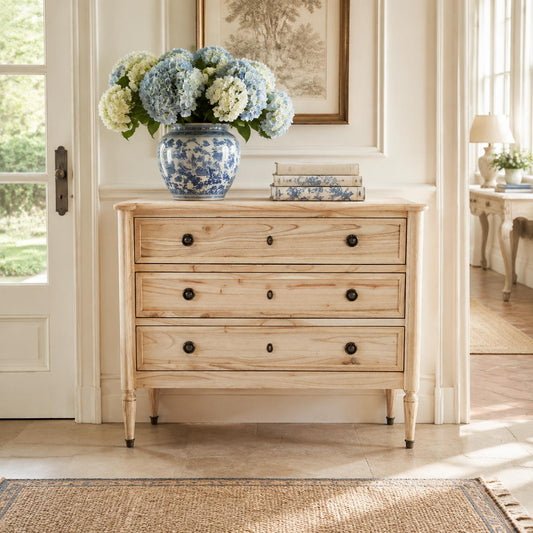 A light natural wood three-drawer chest of drawers with dark round knobs, styled in a bright room with a blue and white ceramic vase of flowers on top.