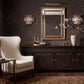 A styled interior scene featuring a dark wood console table beneath a large gold-framed mirror, with a cream upholstered armchair at left, dark patterned wallpaper, wall sconces, and a vase of branches on the tabletop.