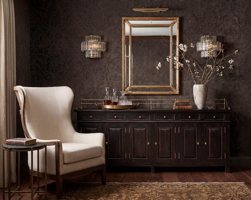 A styled interior scene featuring a dark wood console table beneath a large gold-framed mirror, with a cream upholstered armchair at left, dark patterned wallpaper, wall sconces, and a vase of branches on the tabletop.